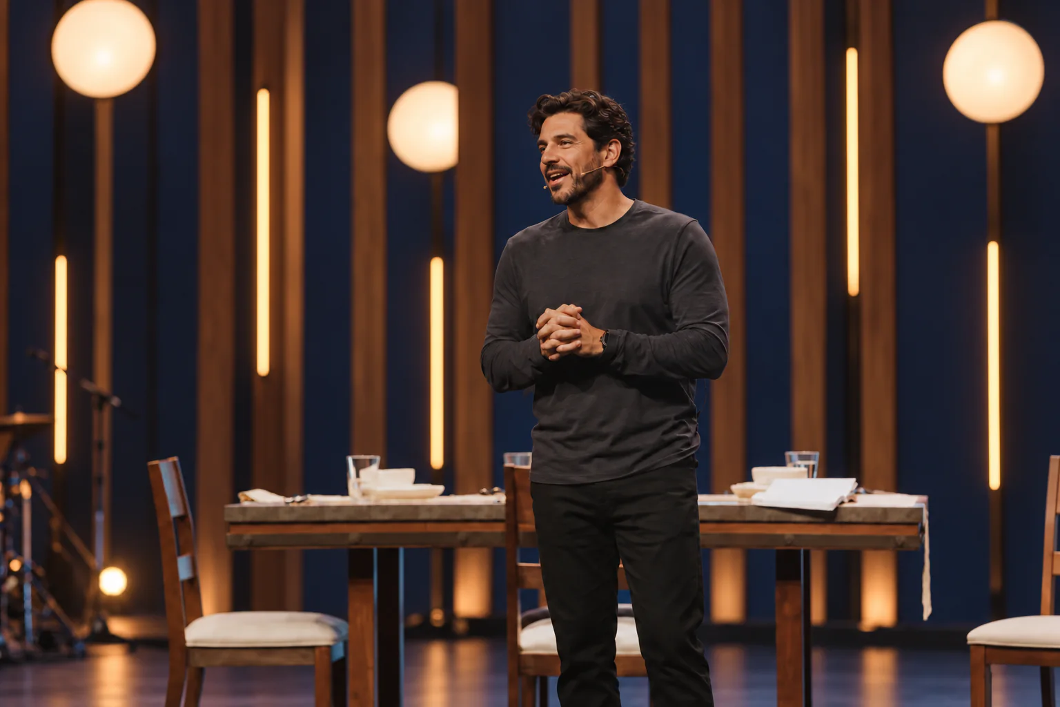 A speaker with a headset microphone talking from a stage set with a long table, wooden slats, and warm globe lights